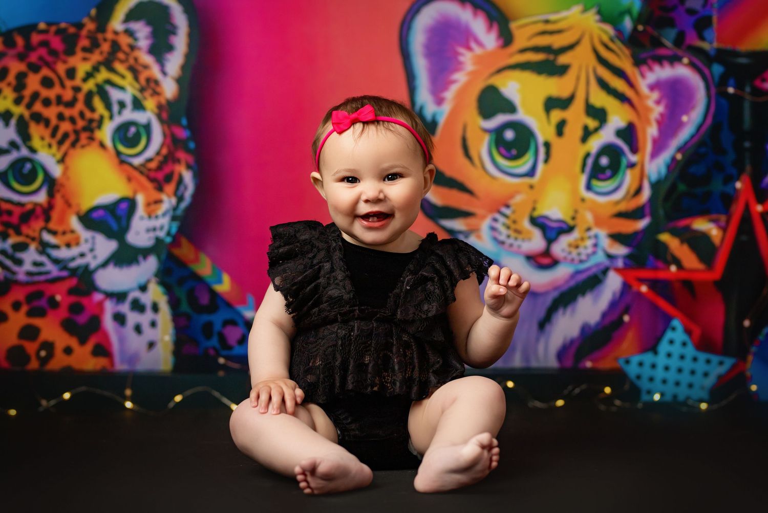 Smiling baby in black romper sits before colorful tiger and leopard backdrop with festive lights.