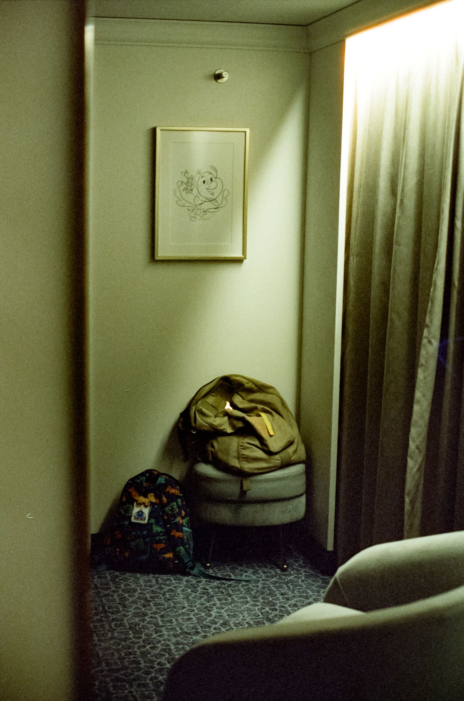 Dimly lit hotel room corner with backpacks and luggage on carpet beneath framed wall art near curtained window.