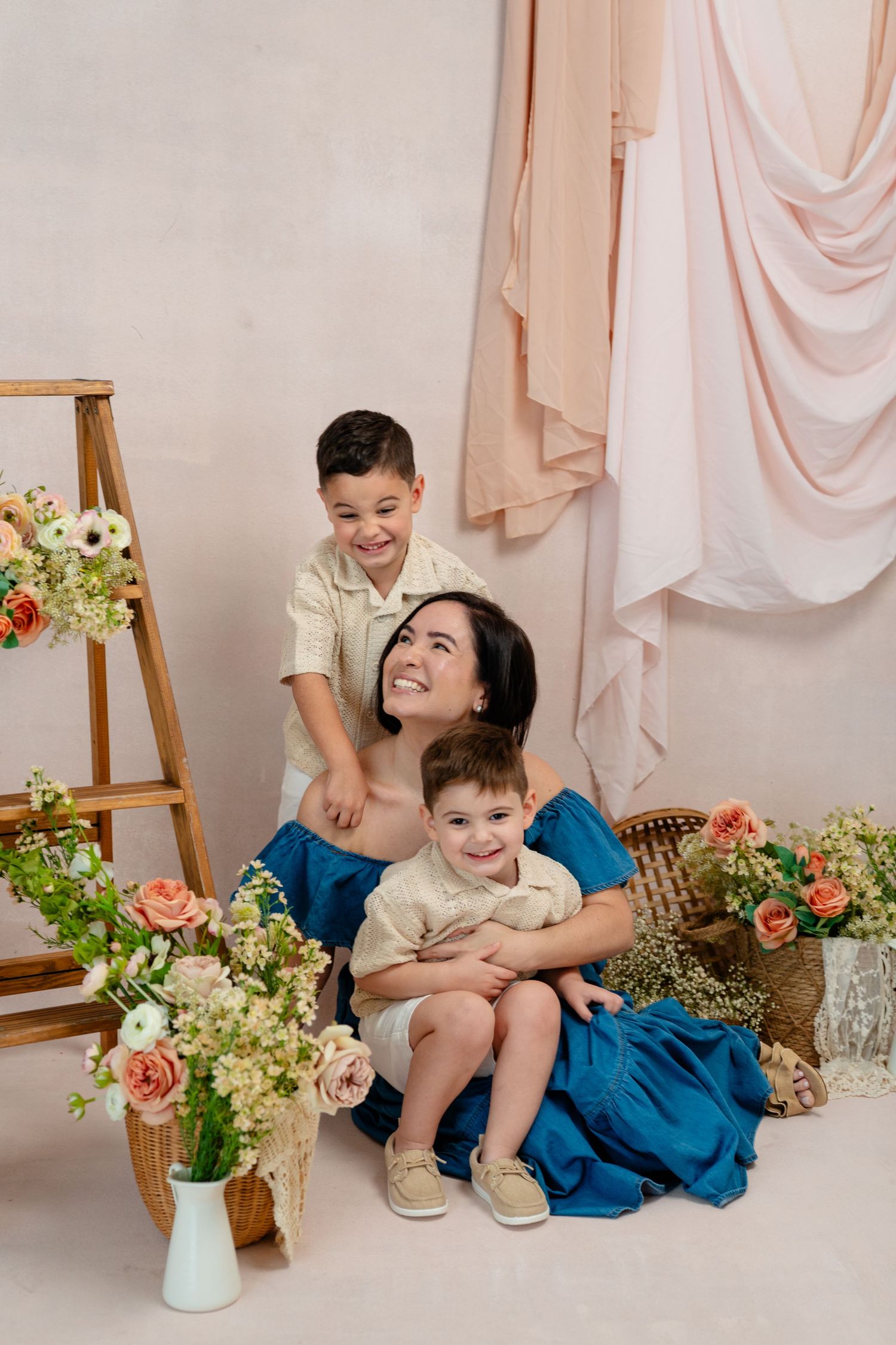 A mother poses with her two young daughters in a floral studio setting, all wearing pastel dresses.