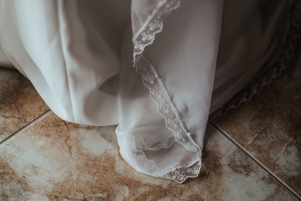 Close-up of a white wedding dress hem with delicate lace trim pooling gracefully on a tiled floor.