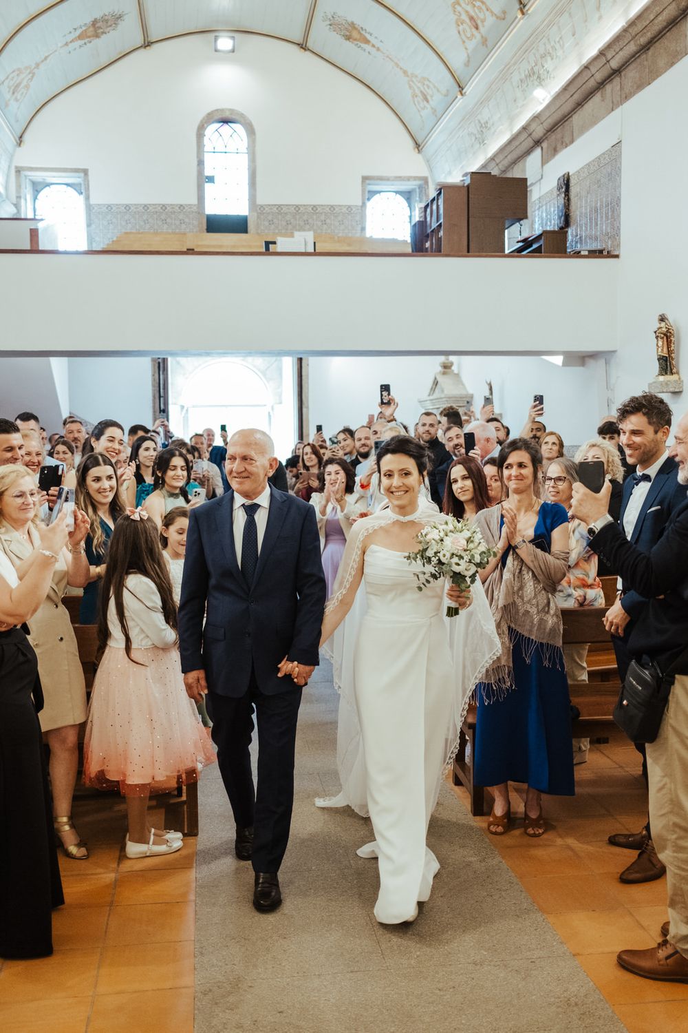 Bride walks down church aisle with escort, guests applauding, holding white bouquet in elegant white gown.