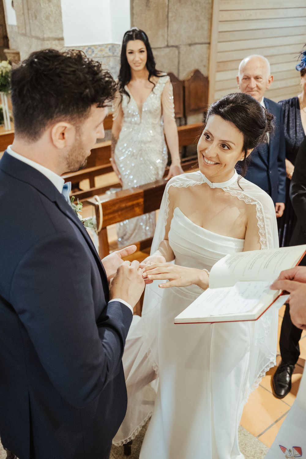 Bride and groom exchange rings during a church wedding ceremony, with guests watching in the background.