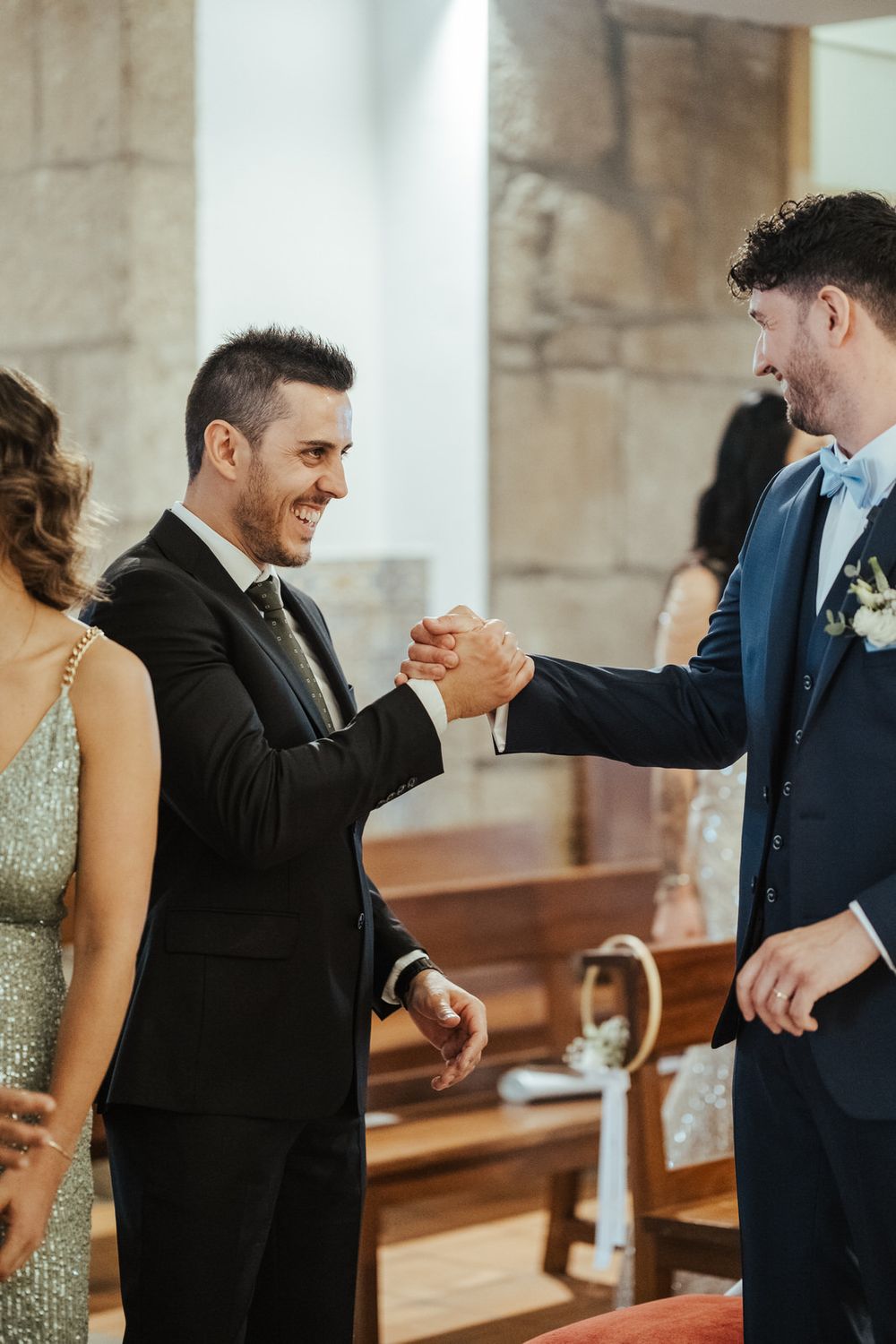 Two smiling men in suits share a warm handshake greeting inside a stone church wedding.