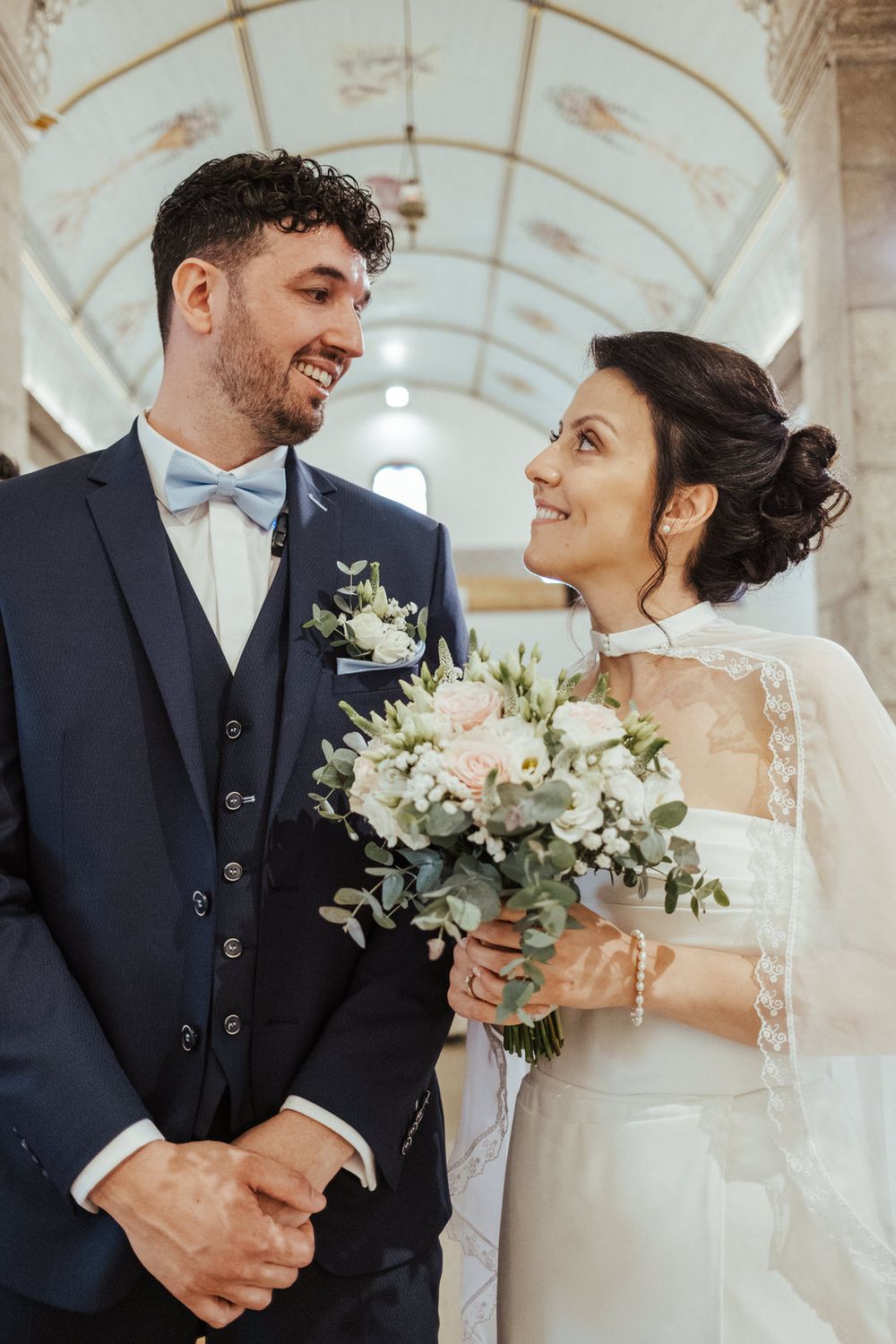 Smiling bride and groom exchange glances inside elegant church, bride holding pastel bouquet, groom in navy suit with blue bow tie.
