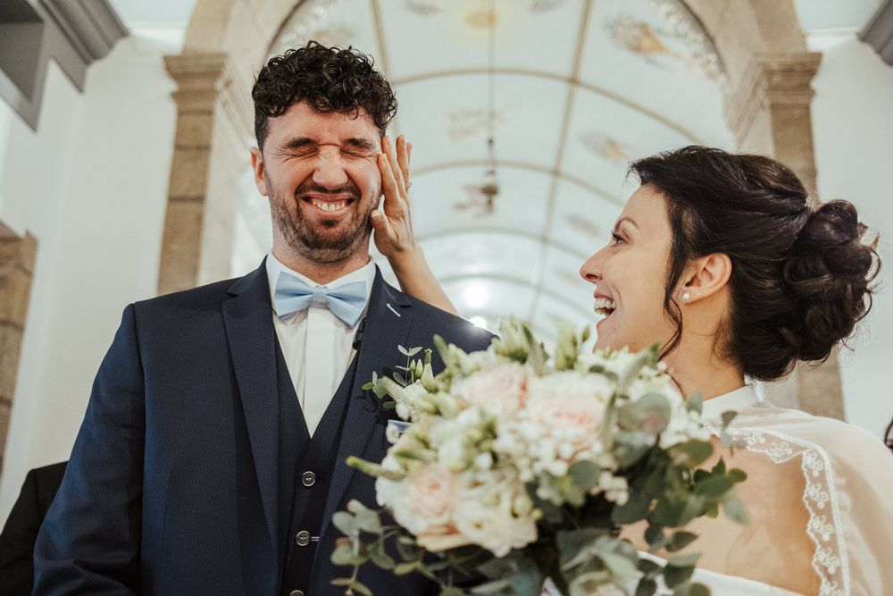 Joyful newlyweds laughing together, groom in navy suit with blue bow tie, bride holding elegant bouquet.