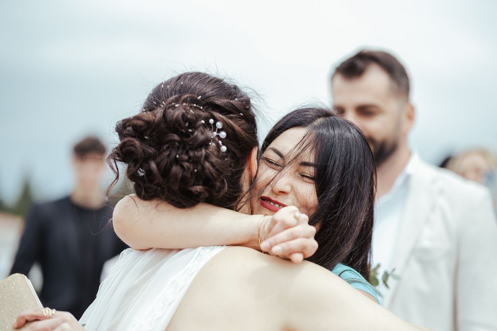 A bride and her guest share an emotional hug during an outdoor wedding ceremony, with a groom visible in the background.