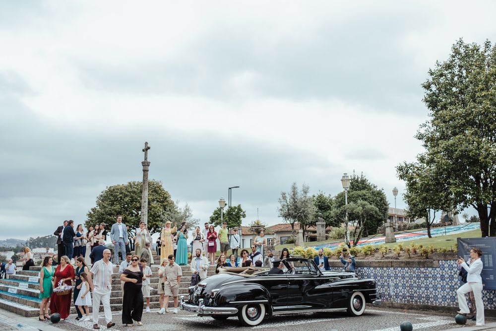 Vintage black classic car at an outdoor gathering with guests near a monument by the waterfront on a cloudy day.