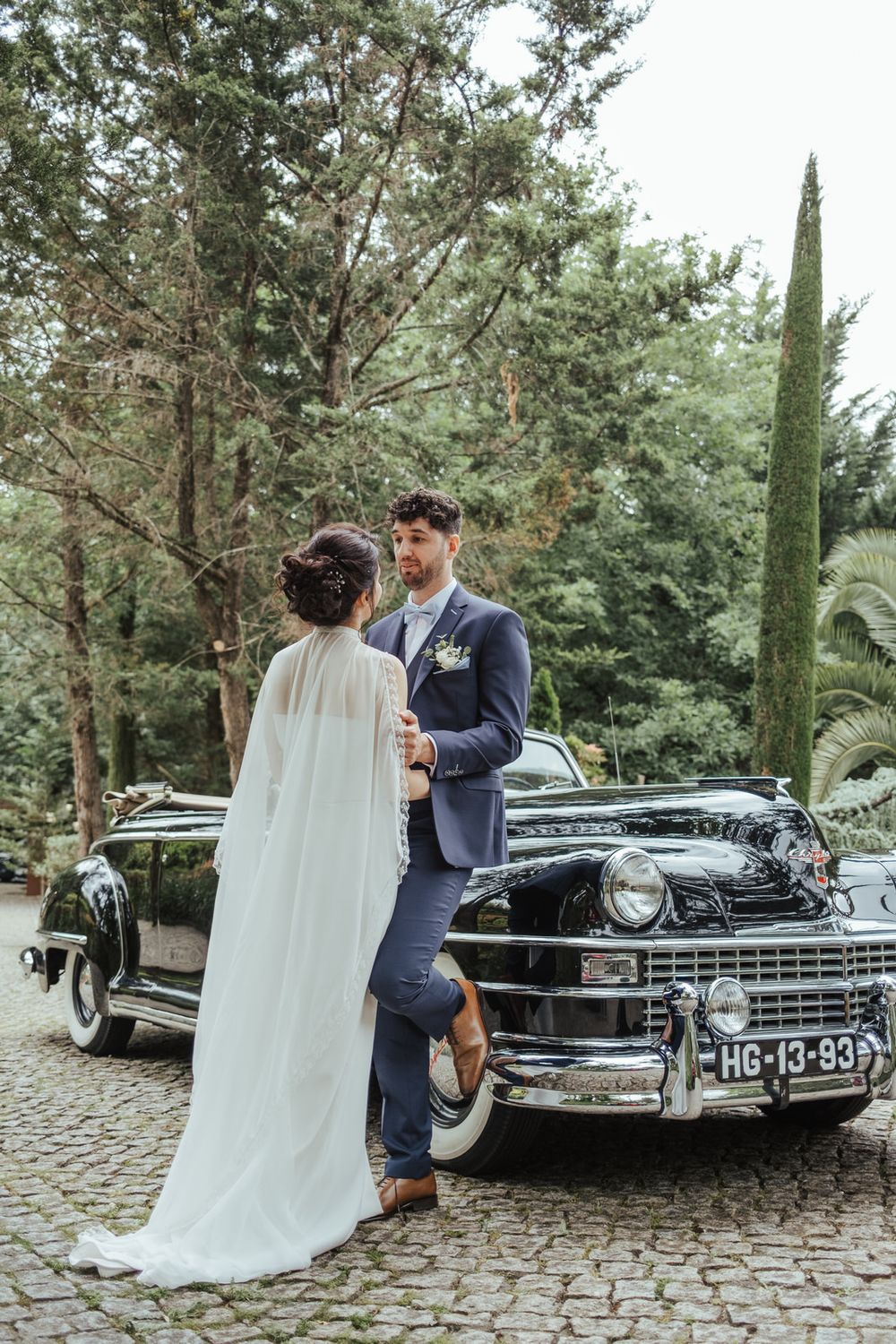 Bride and groom posing beside a vintage black classic car on cobblestone, surrounded by lush green trees.
