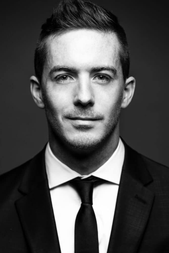 Black and white professional portrait of a young man in a suit and tie against a dark background.