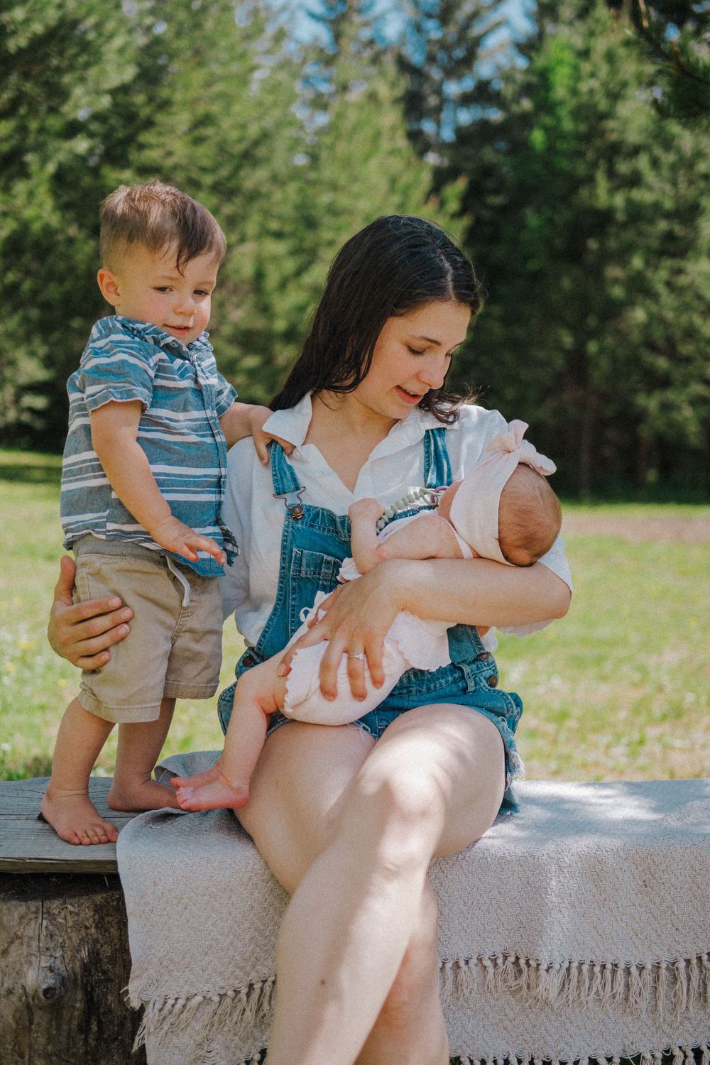 A mother sits outdoors holding a newborn while a toddler boy stands beside her in a sunny green park.
