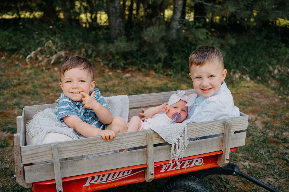 Two young boys and a newborn baby sitting together in a red Radio Flyer wagon outdoors.