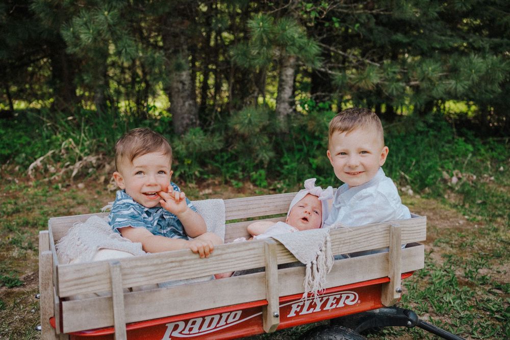 Two toddlers and a newborn baby sitting together in a classic red Radio Flyer wagon outdoors.