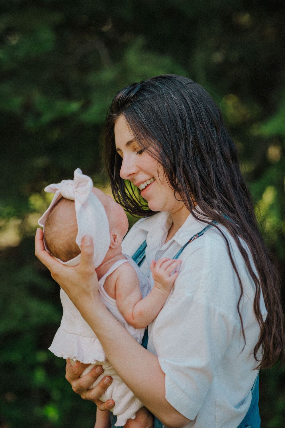 Smiling young mother with long dark hair holding her newborn baby outdoors among green trees.