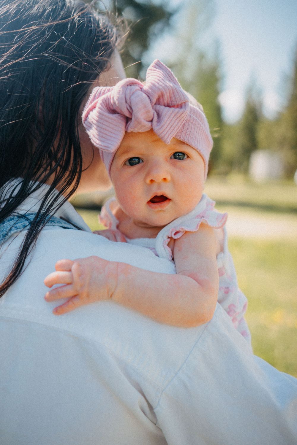 A baby girl in a pink bow headband and floral dress being held over a mother's shoulder outdoors.