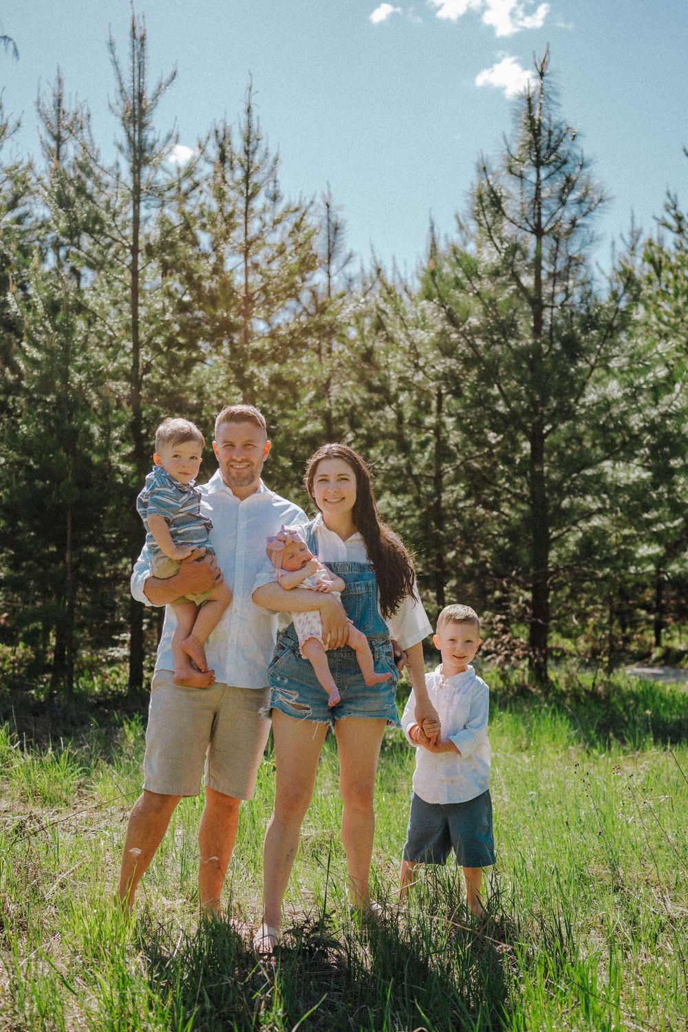 Happy family of five posing outdoors in a sunny pine forest, parents holding young children on a bright summer day.