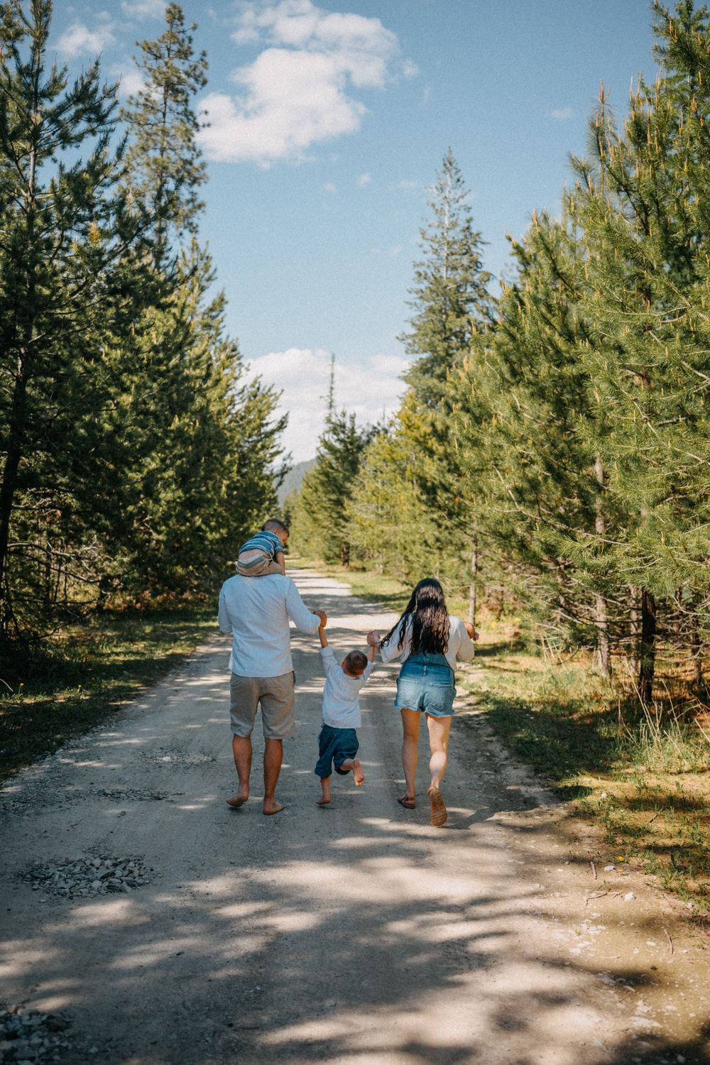 A family of three walks barefoot down a sunny forest path, holding hands with their toddler.