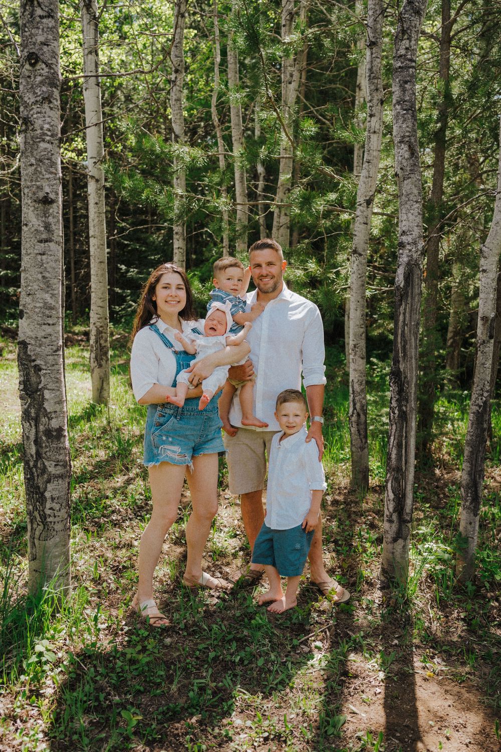 Happy family of four posing in a sunlit birch forest, parents holding young children, wearing casual summer outfits.