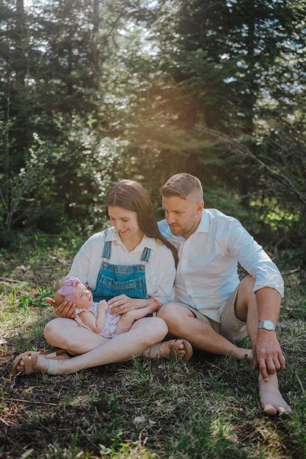 Family sitting on grass in sunlit forest, mother holding baby girl in pink outfit while father looks on lovingly.