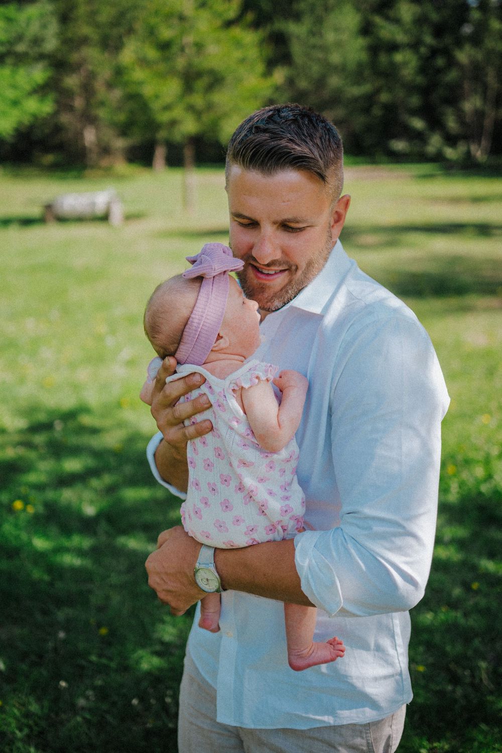 A smiling father in a white shirt lovingly holds his newborn baby girl in a sunny green park.