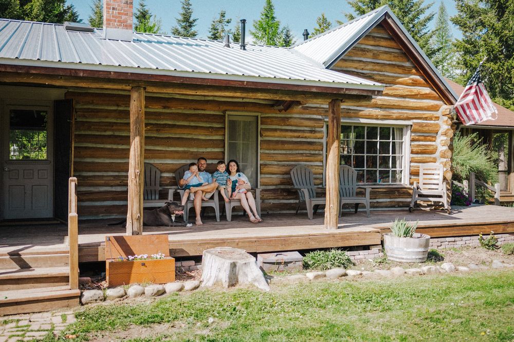 Family relaxing on the porch of a rustic log cabin with metal roof, surrounded by trees and an American flag.
