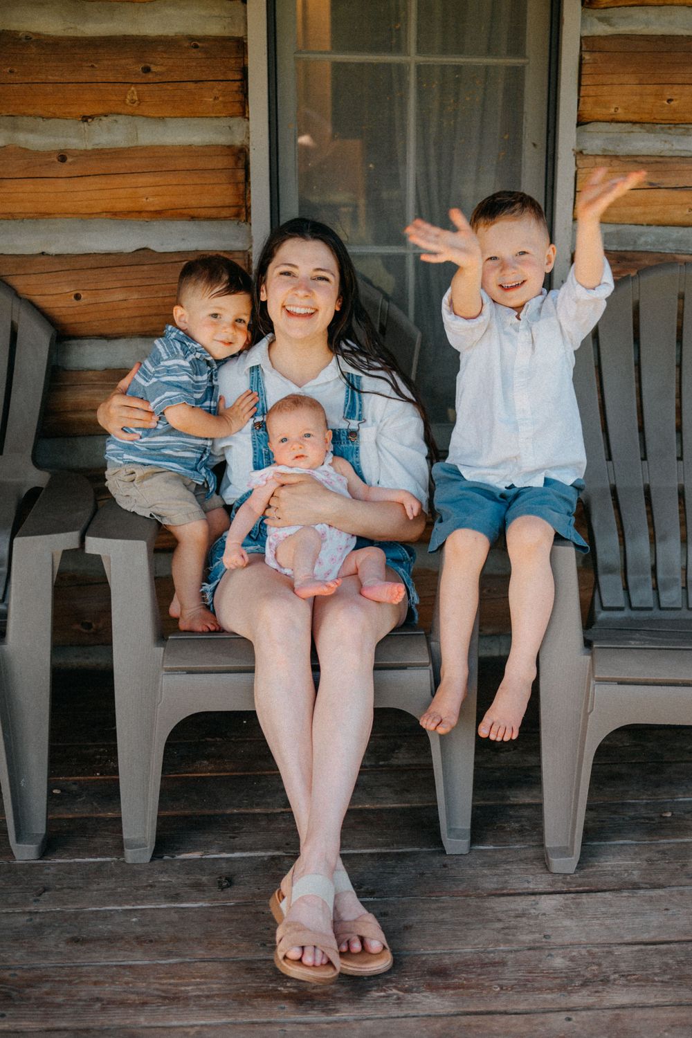 A smiling mother sits on a porch with three young children, including a baby on her lap, beside a rustic log cabin.