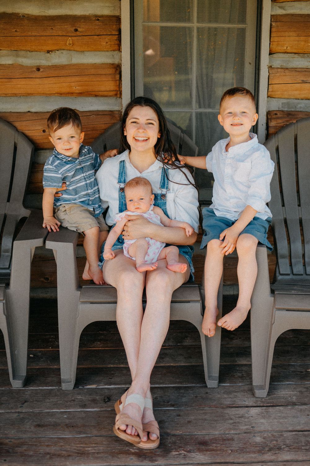 A smiling mother sits on a porch with three young children, including a baby, in front of a rustic log cabin.