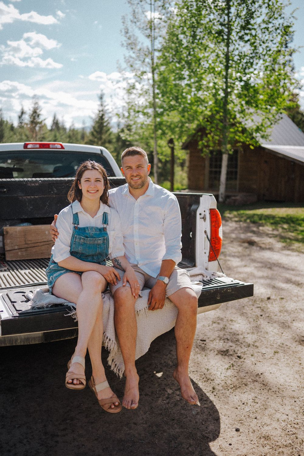 Couple sitting together on a pickup truck tailgate outdoors near a cabin surrounded by trees on a sunny day.