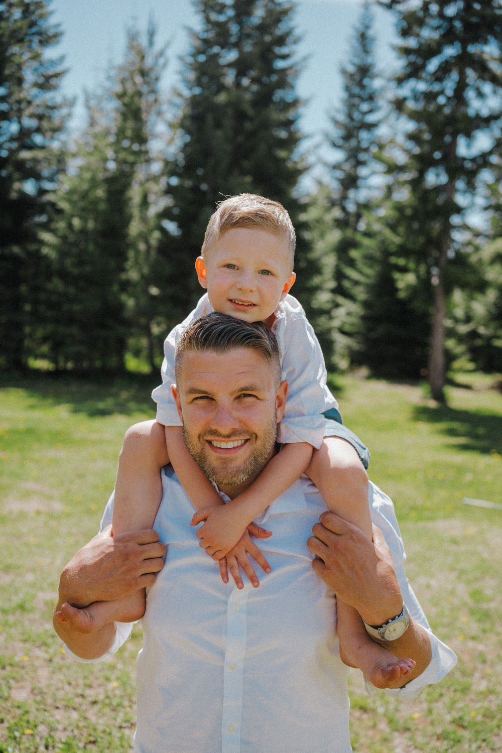 Smiling man carries young boy on his shoulders in a sunny green park with tall pine trees in the background.