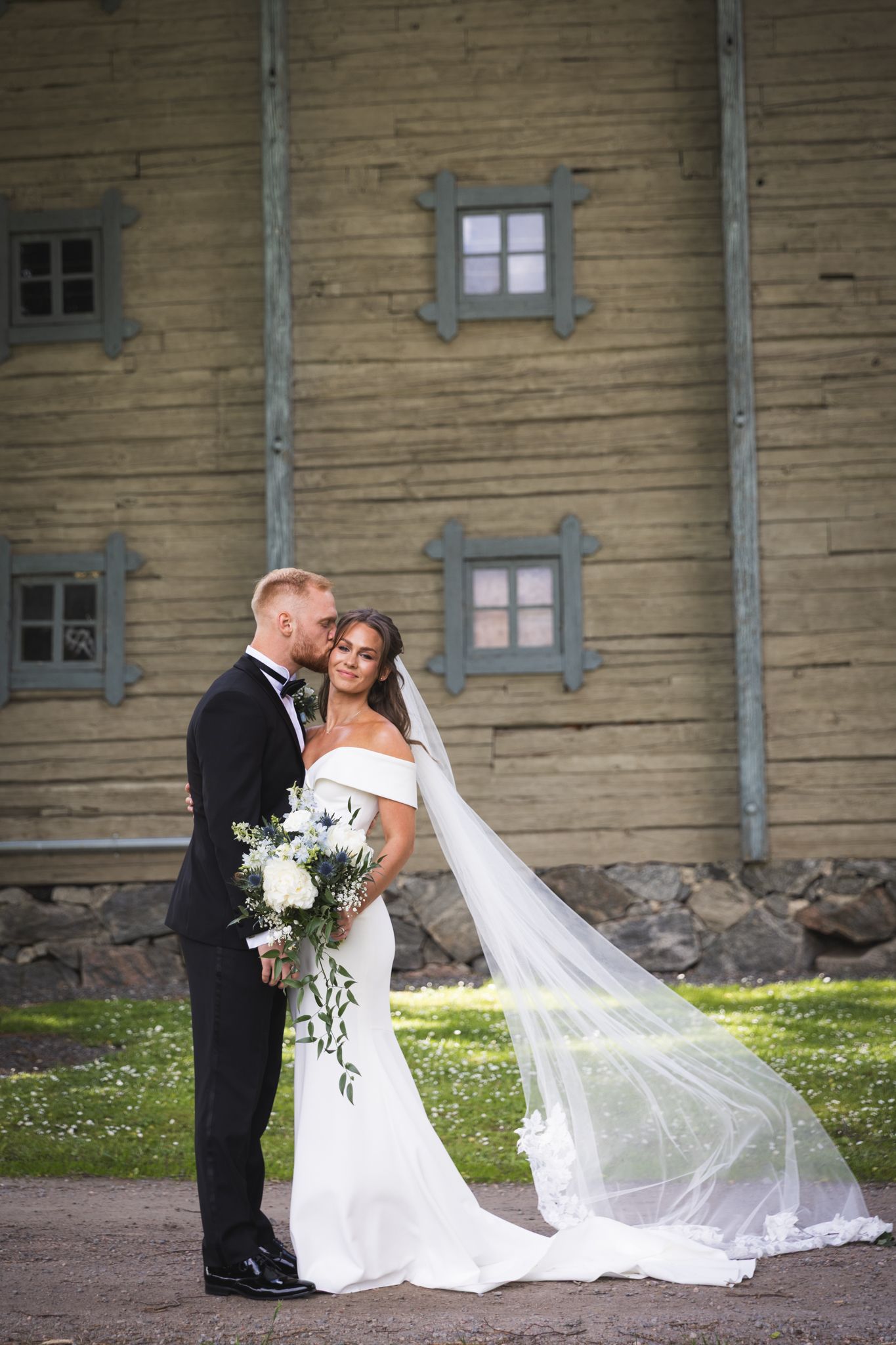 A couple shares a romantic moment in their wedding attire against a rustic wooden building with small windows.