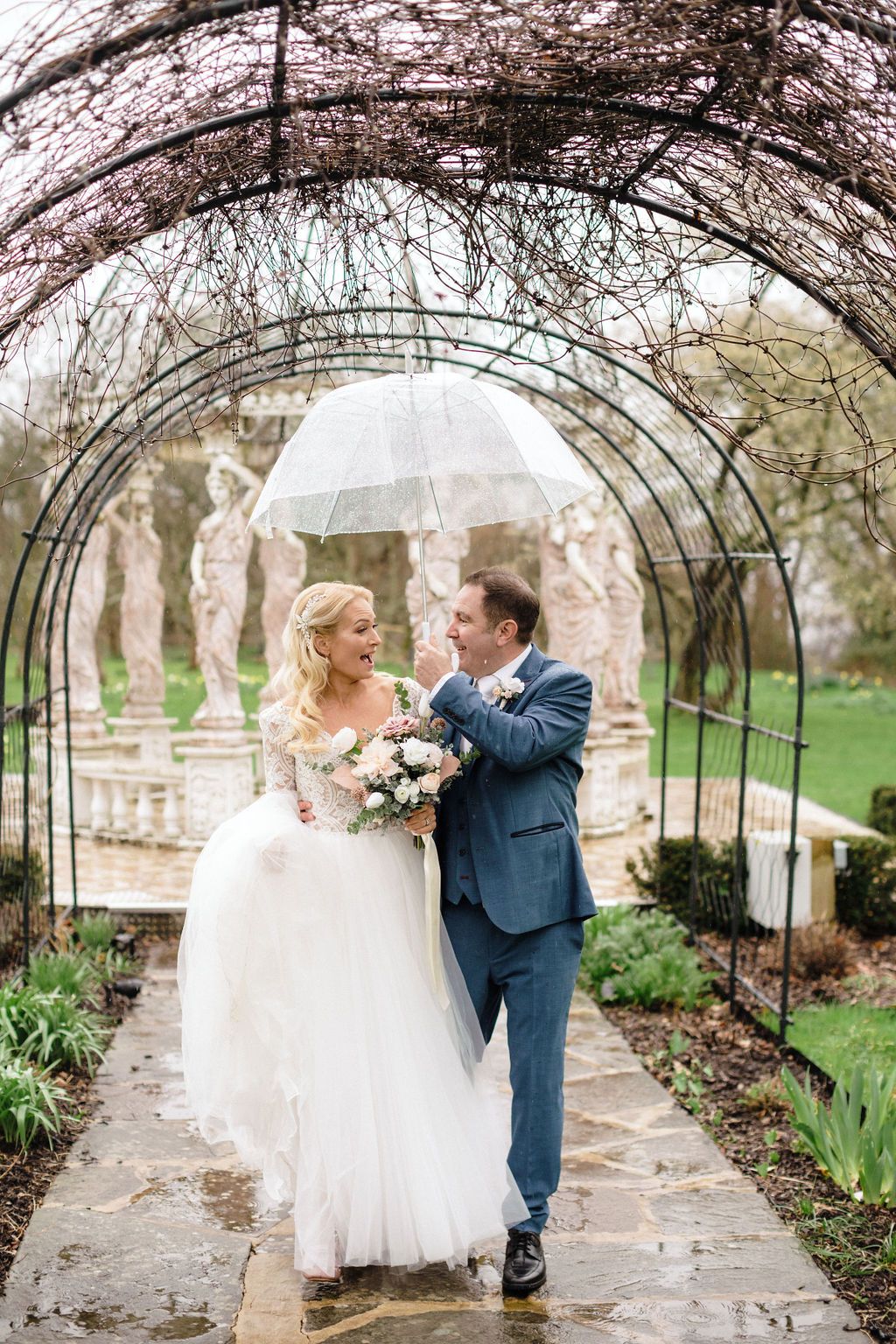 Groom carries bride under blue umbrella in rain at outdoor wedding ceremony.