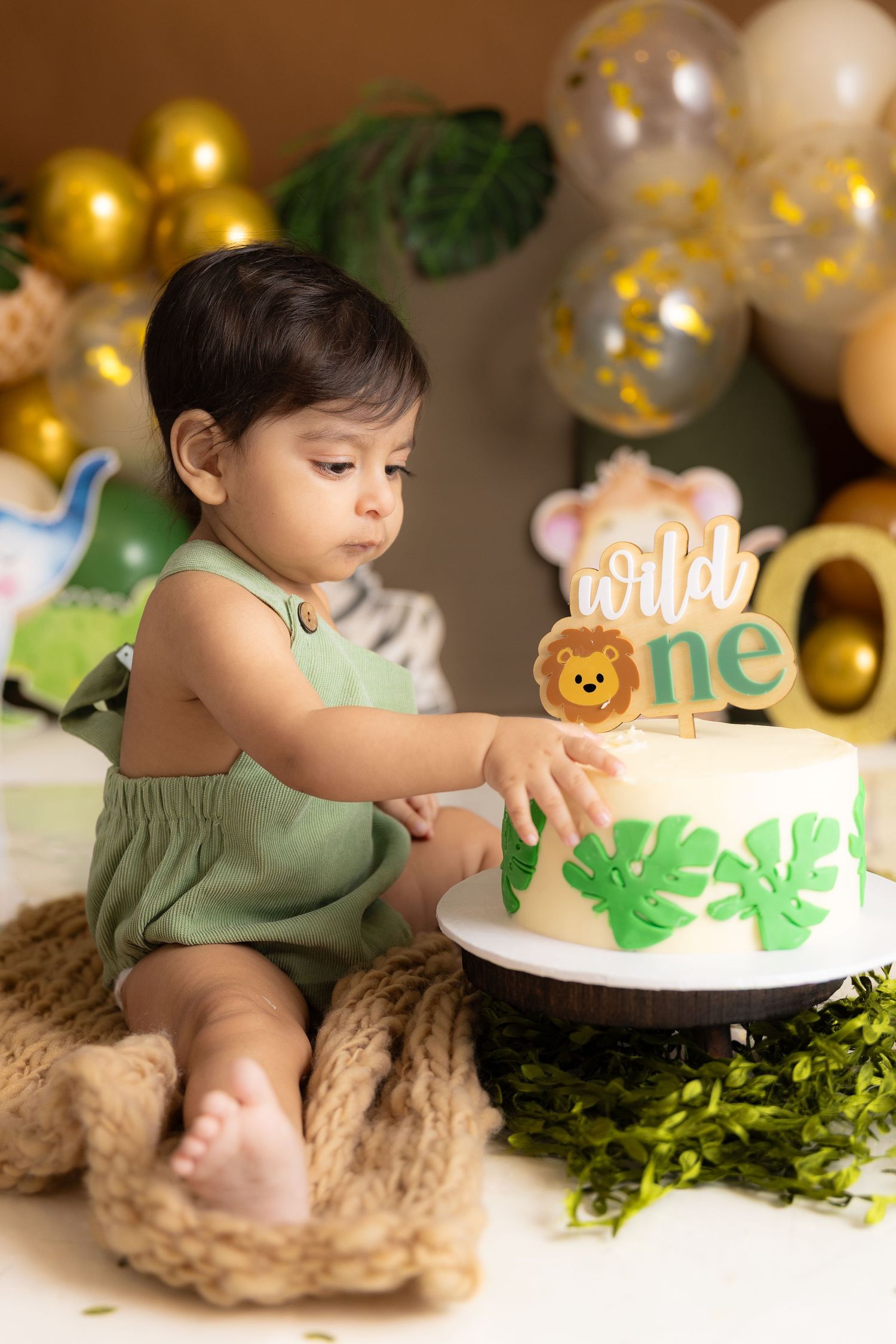 A baby boy in a green romper reaches toward a Wild One safari-themed first birthday cake surrounded by gold and green balloons.