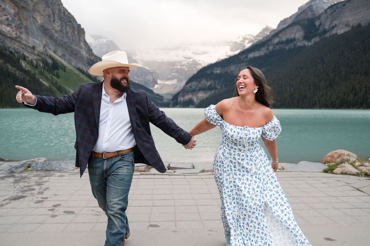 Couple laughing and dancing hand-in-hand at Lake Louise with turquoise water and Rocky Mountains backdrop.