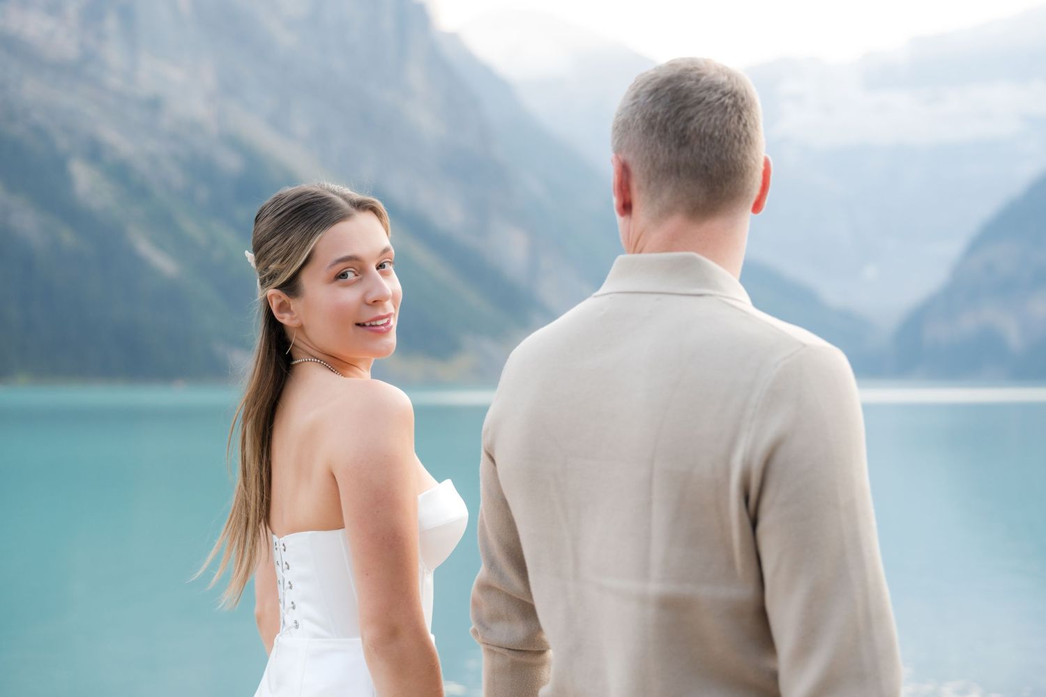 Woman in white dress smiles at fiance in tan sweater by turquoise Lake Louise, Banff during surprise proposal photoshoot