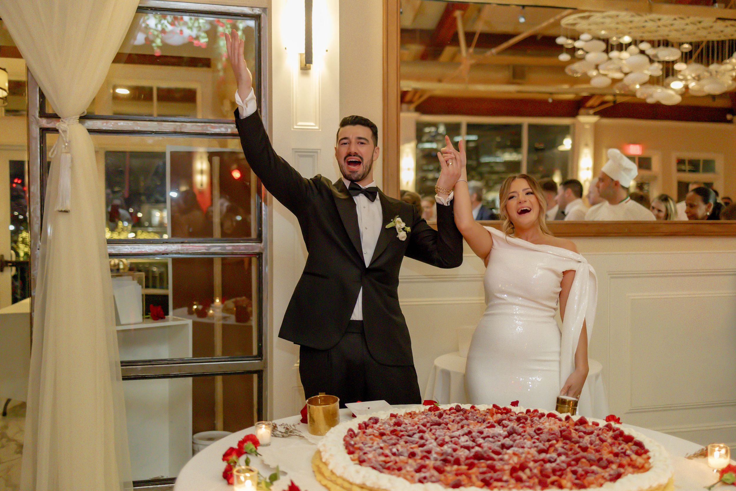 Joyful bride and groom celebrating at wedding reception beside a stunning strawberry-topped cake at Liberty House in Jersey City, NJ.