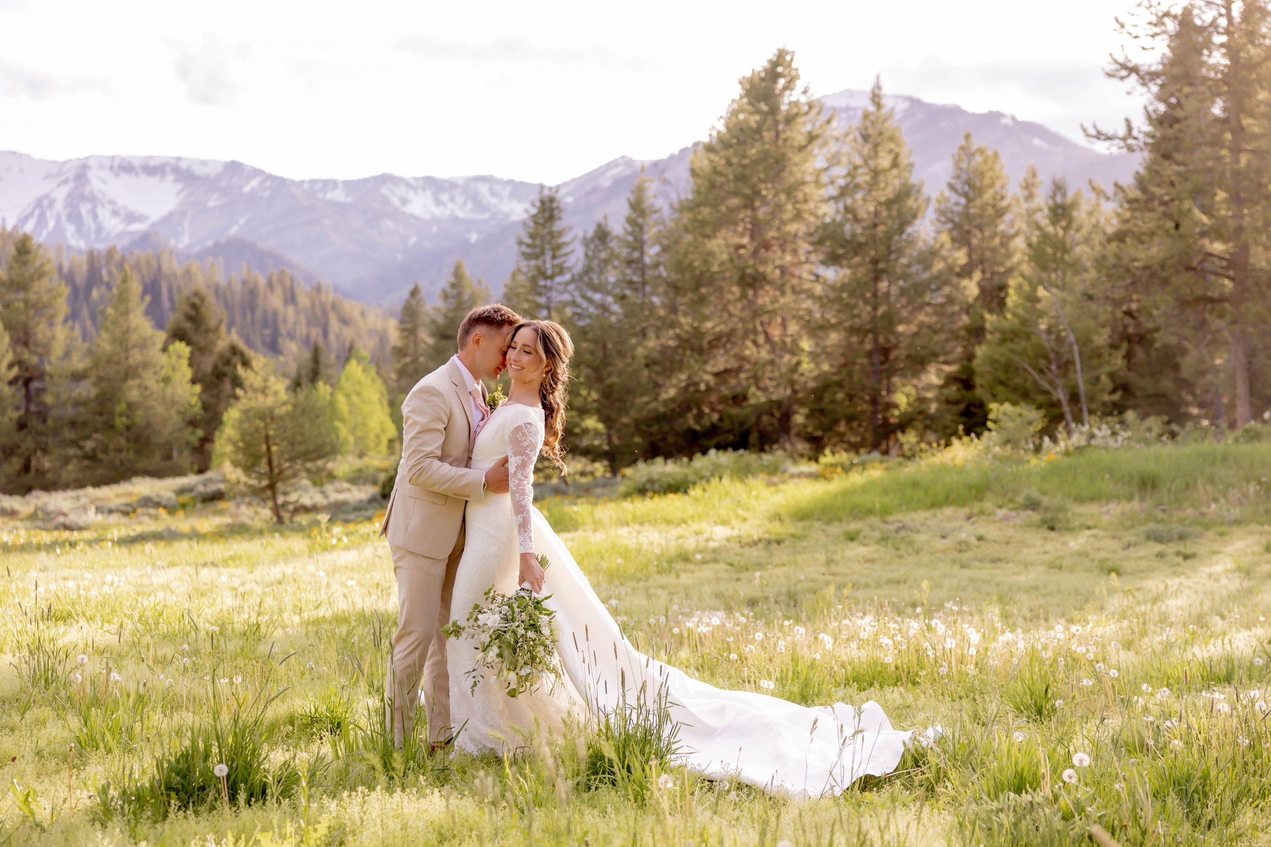Romantic bride and groom embracing in sunlit mountain meadow with snow-capped peaks behind them in Jackson, Wyoming.