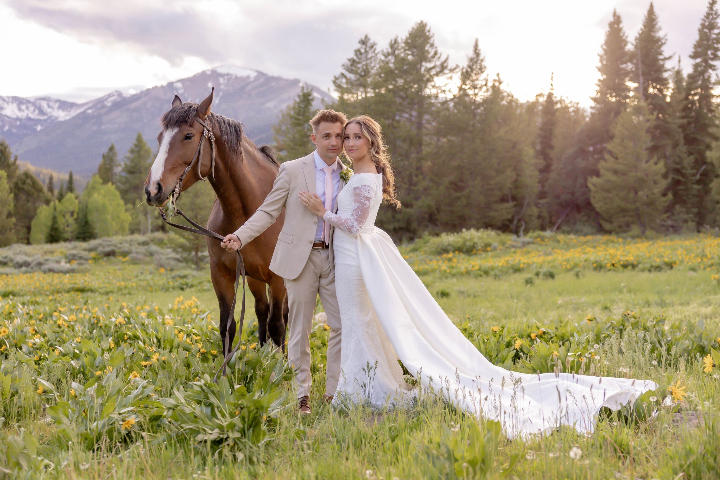 Bride and groom posing with brown horse in scenic mountain meadow with snow-capped peaks backdrop in Jackson, Wyoming.