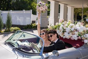 Joyful bride raises bouquet while riding with groom in flower-adorned vintage convertible on sunny day.