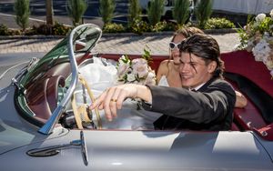 Smiling groom looks back at bride in flower-decorated vintage convertible car on wedding day.
