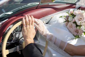 Close-up of bride and groom holding hands on steering wheel of classic convertible with pink bouquet.