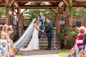 Bride and groom exchange vows under a wooden gazebo adorned with colorful flowers during outdoor ceremony at Tourelay Farm in Colts Neck, NJ