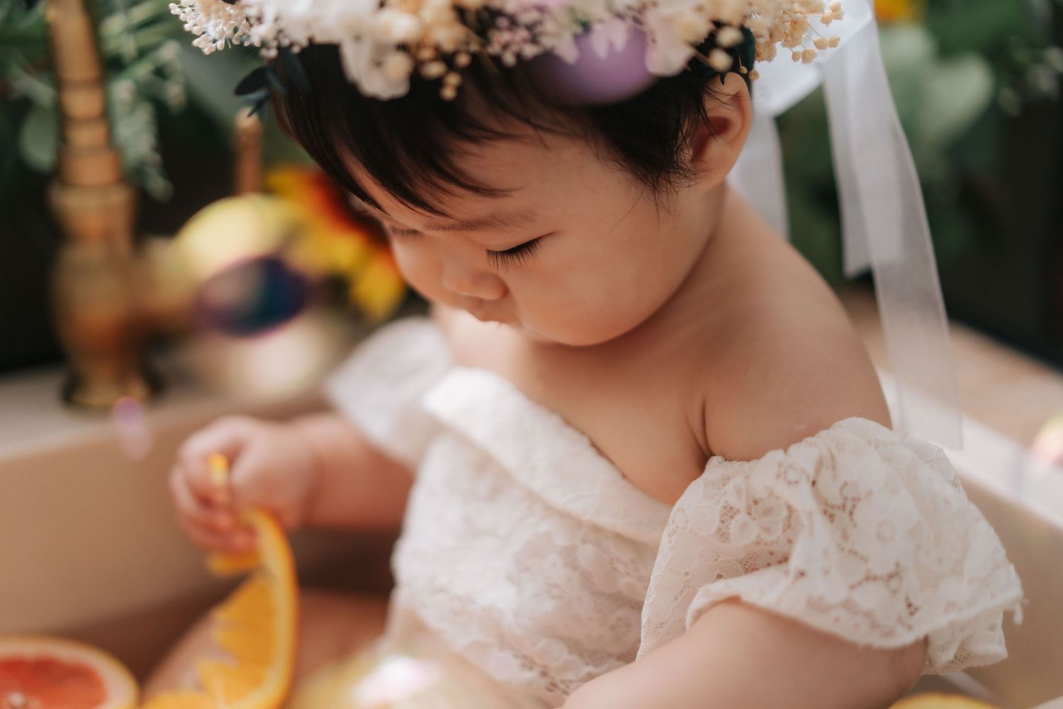 A child wearing a delicate white lace dress and floral crown looks down while holding something yellow.一個穿著精緻白色蕾絲洋裝、頭戴花冠的孩子低著頭,手裡拿著一個黃色的東西。