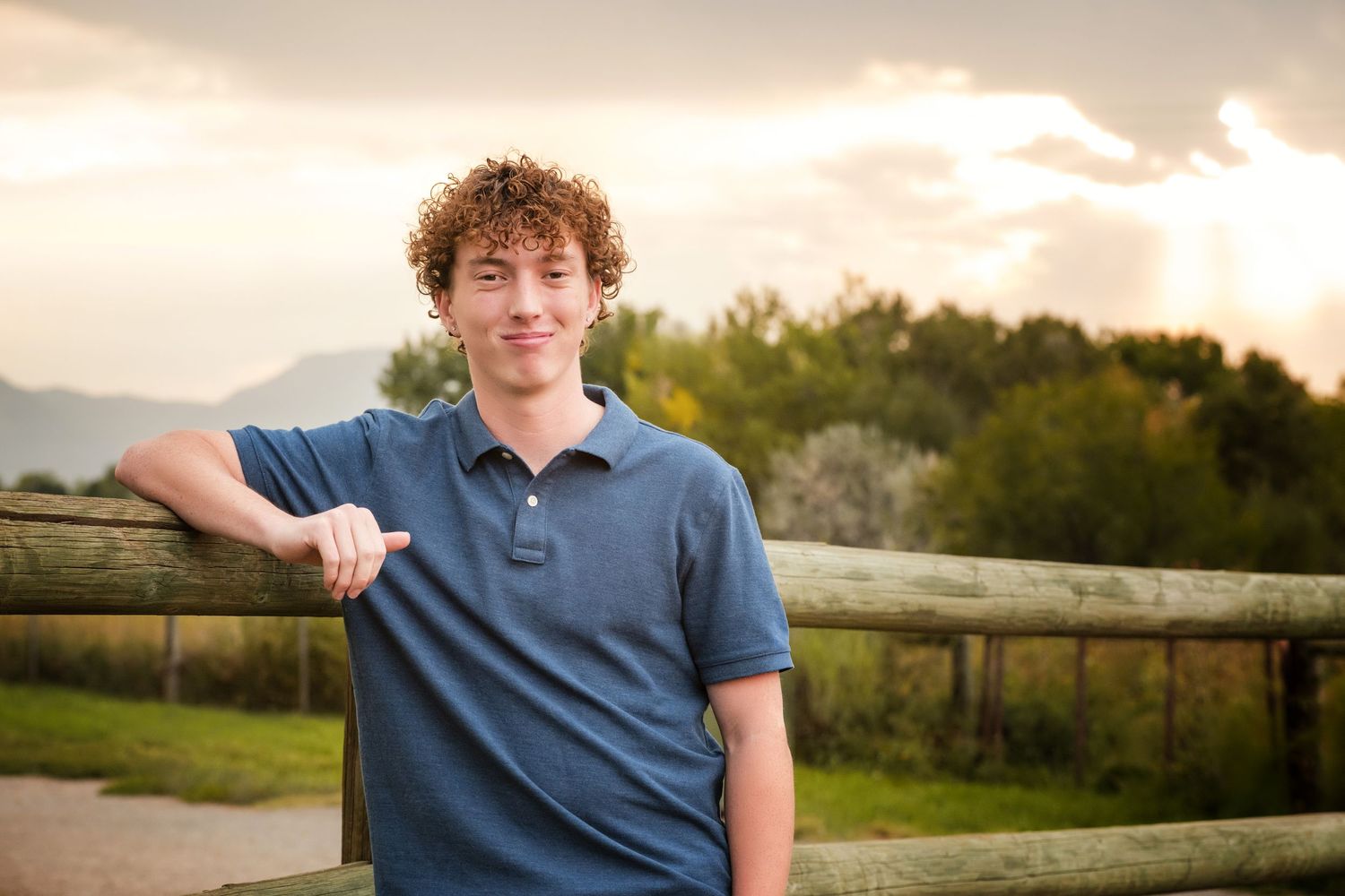 Smiling young man with curly hair giving thumbs up near wooden fence with scenic mountain backdrop.