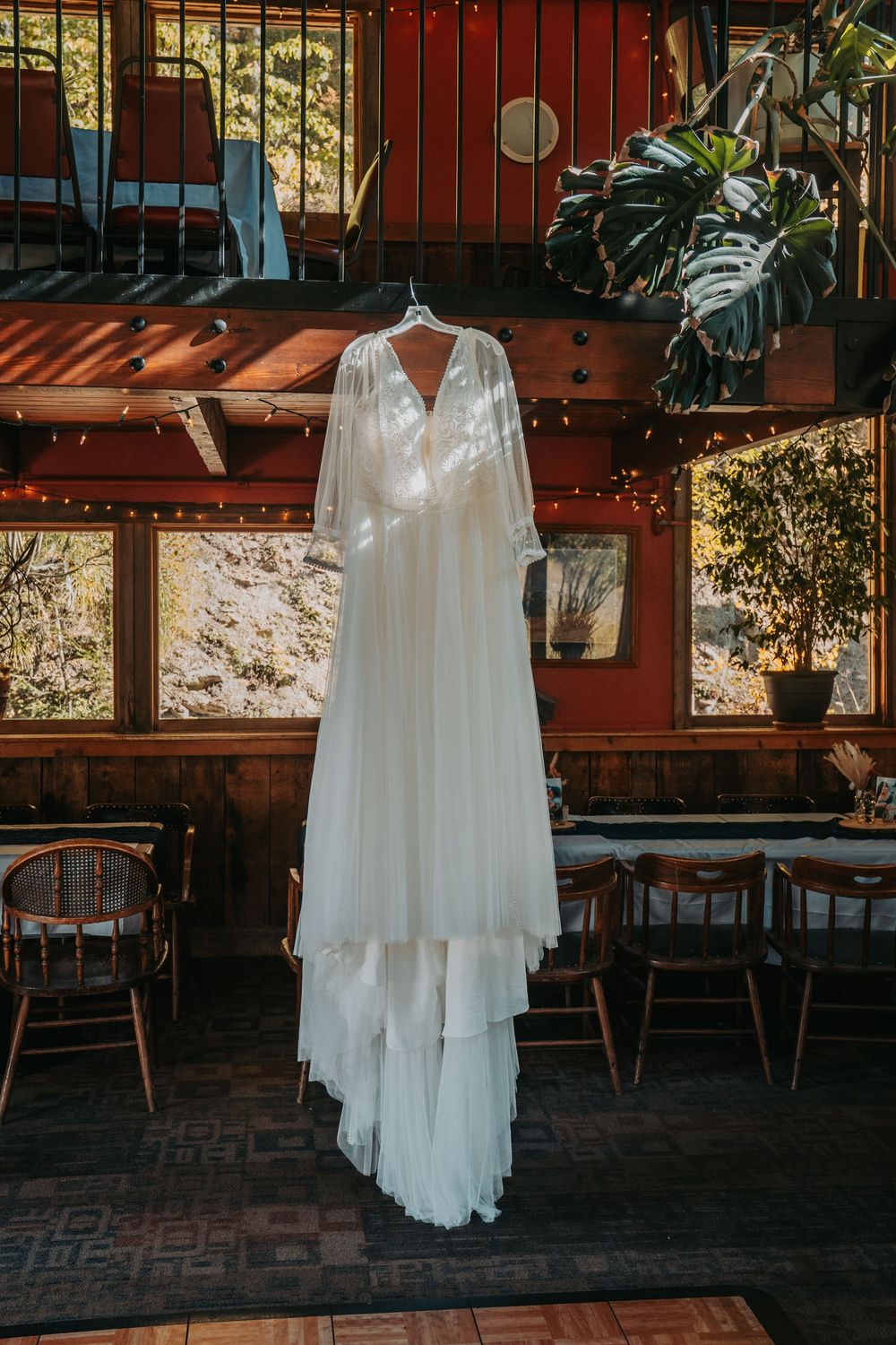 Elegant white wedding dress hanging in a rustic wooden venue with string lights and large windows.
