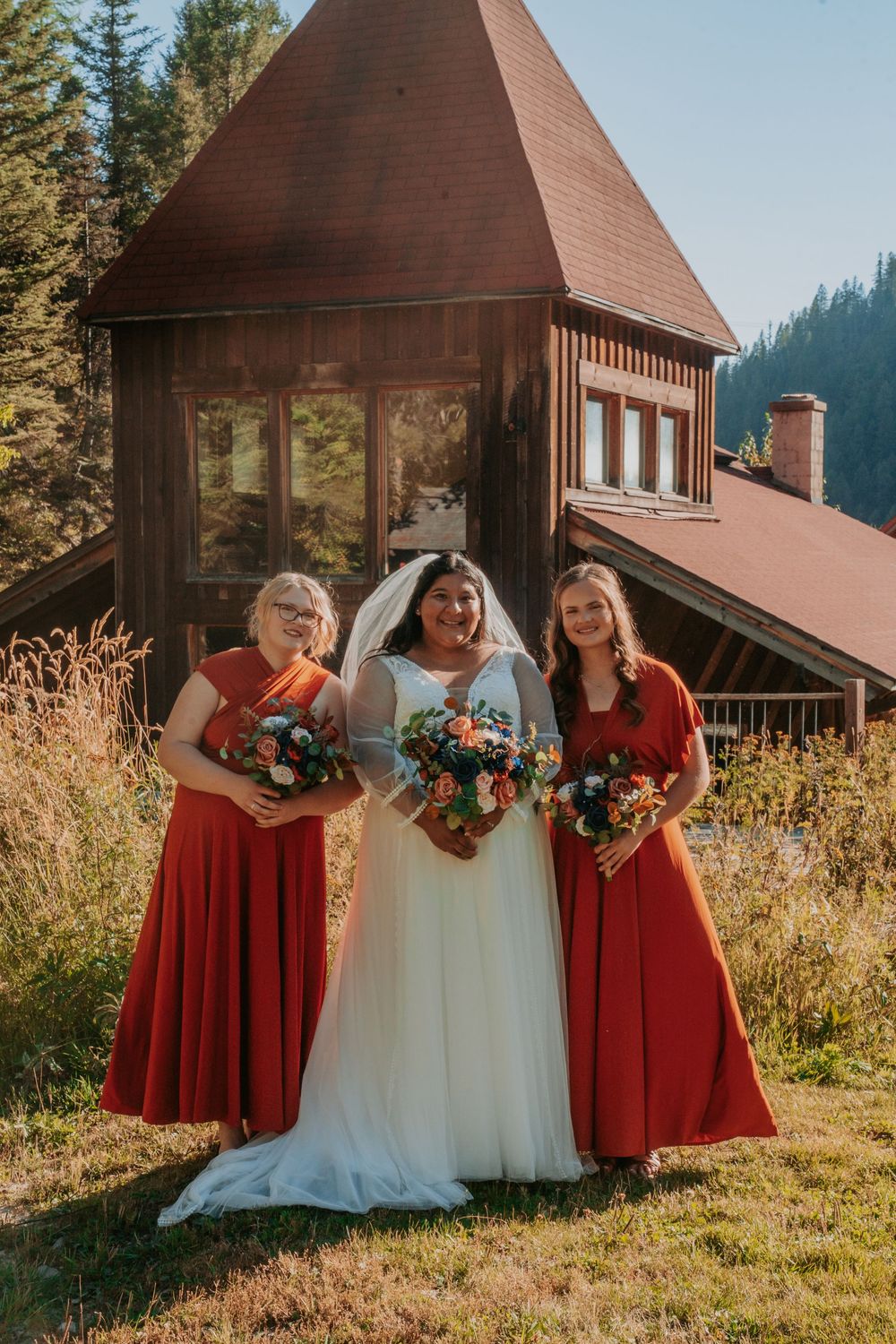 Bride in white gown poses with two bridesmaids in rust red dresses holding bouquets outside a rustic wooden chapel.
