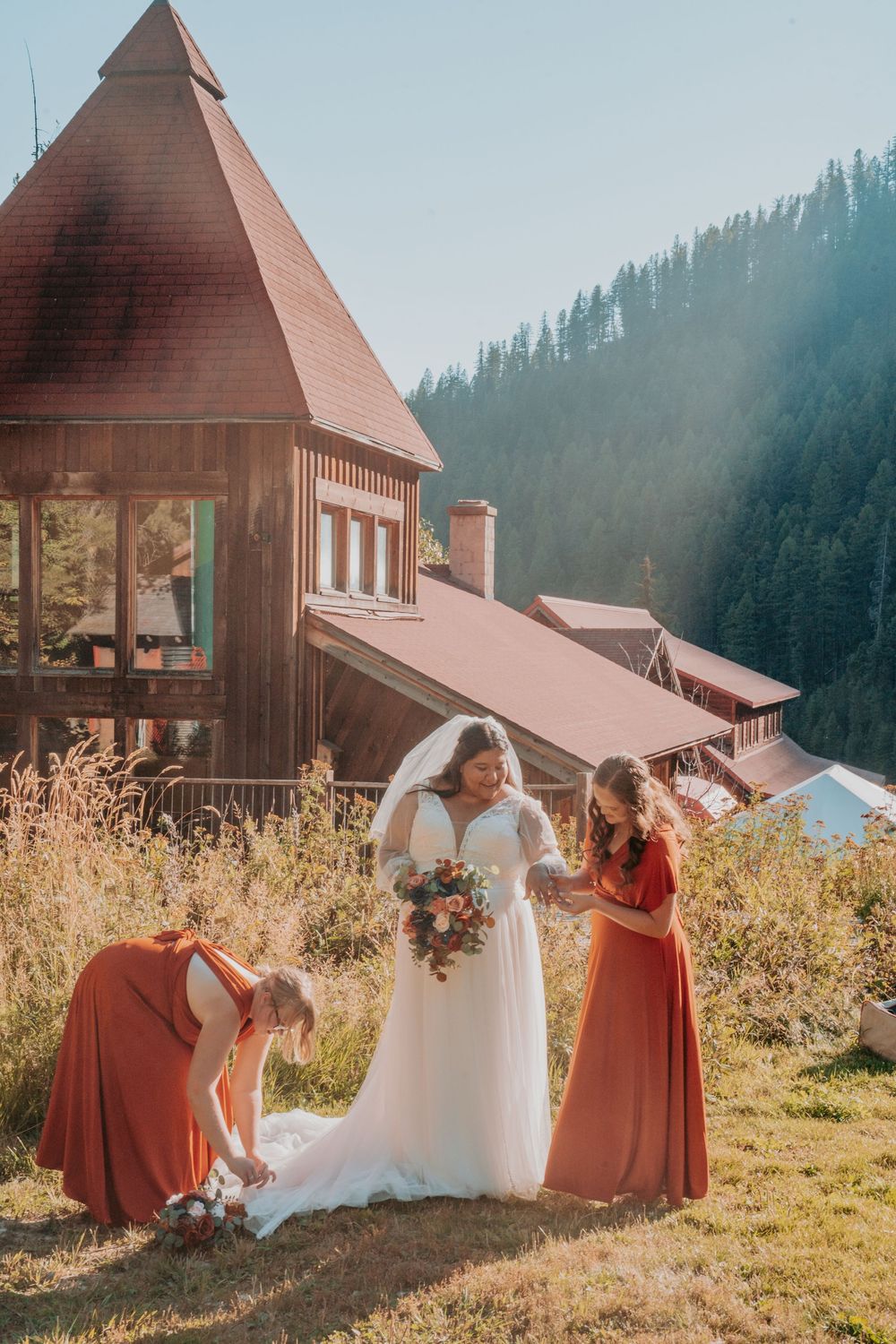 Bride in white gown with bridesmaids in rust dresses outdoors near rustic mountain lodge on sunny day.