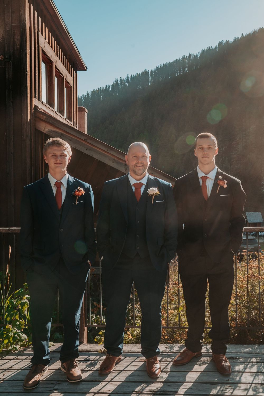 Three groomsmen in navy suits with floral boutonnieres posing outdoors on a sunny mountain wedding day.