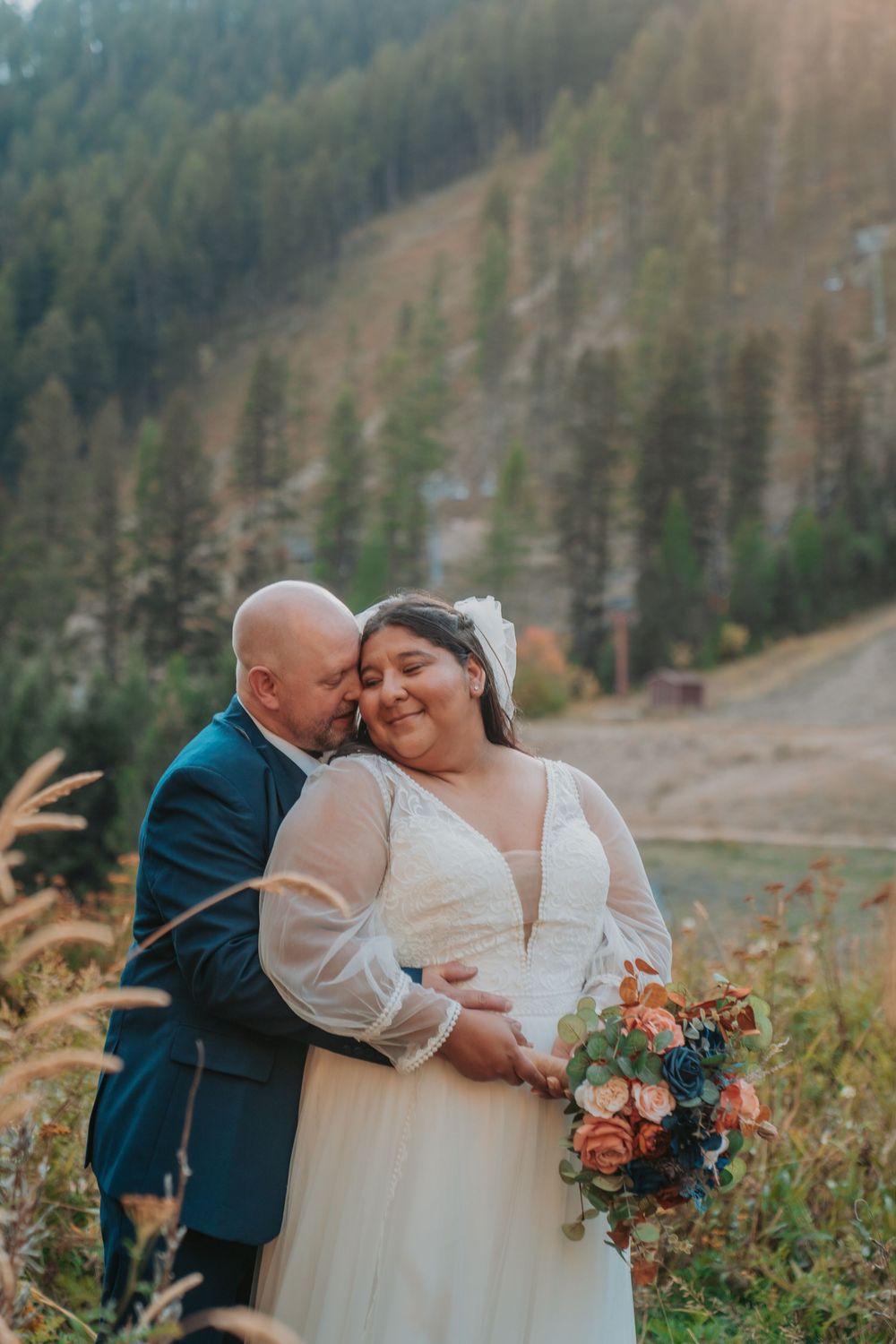 Groom in navy suit embraces bride in white lace gown holding a blue and coral bouquet in a mountain setting.