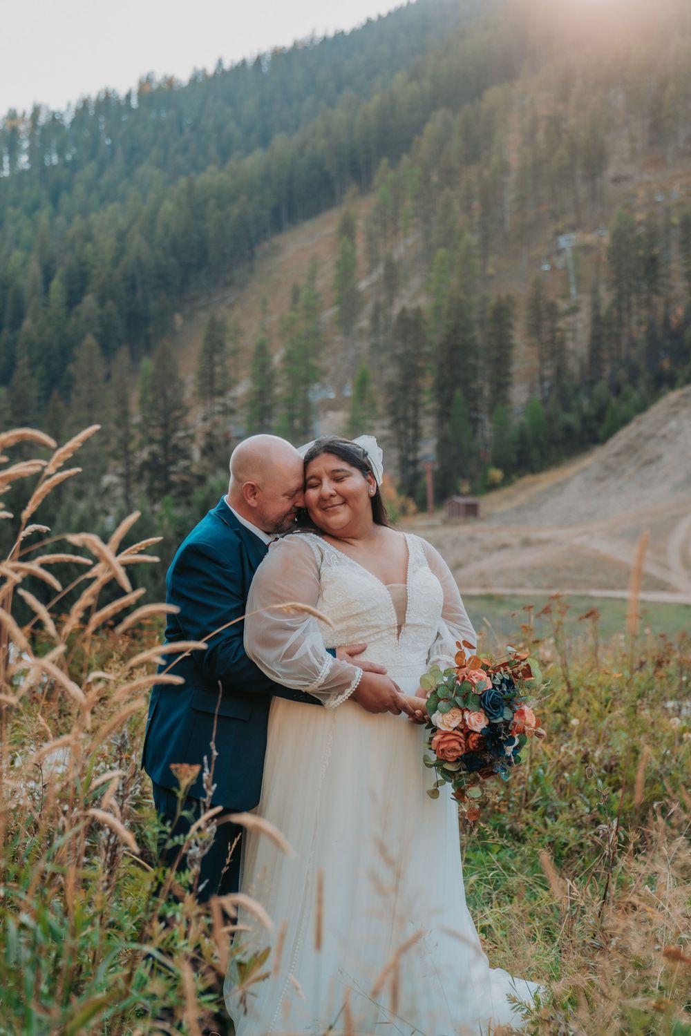 Bride and groom embrace in a golden field with pine-covered mountains behind them during an outdoor fall wedding.