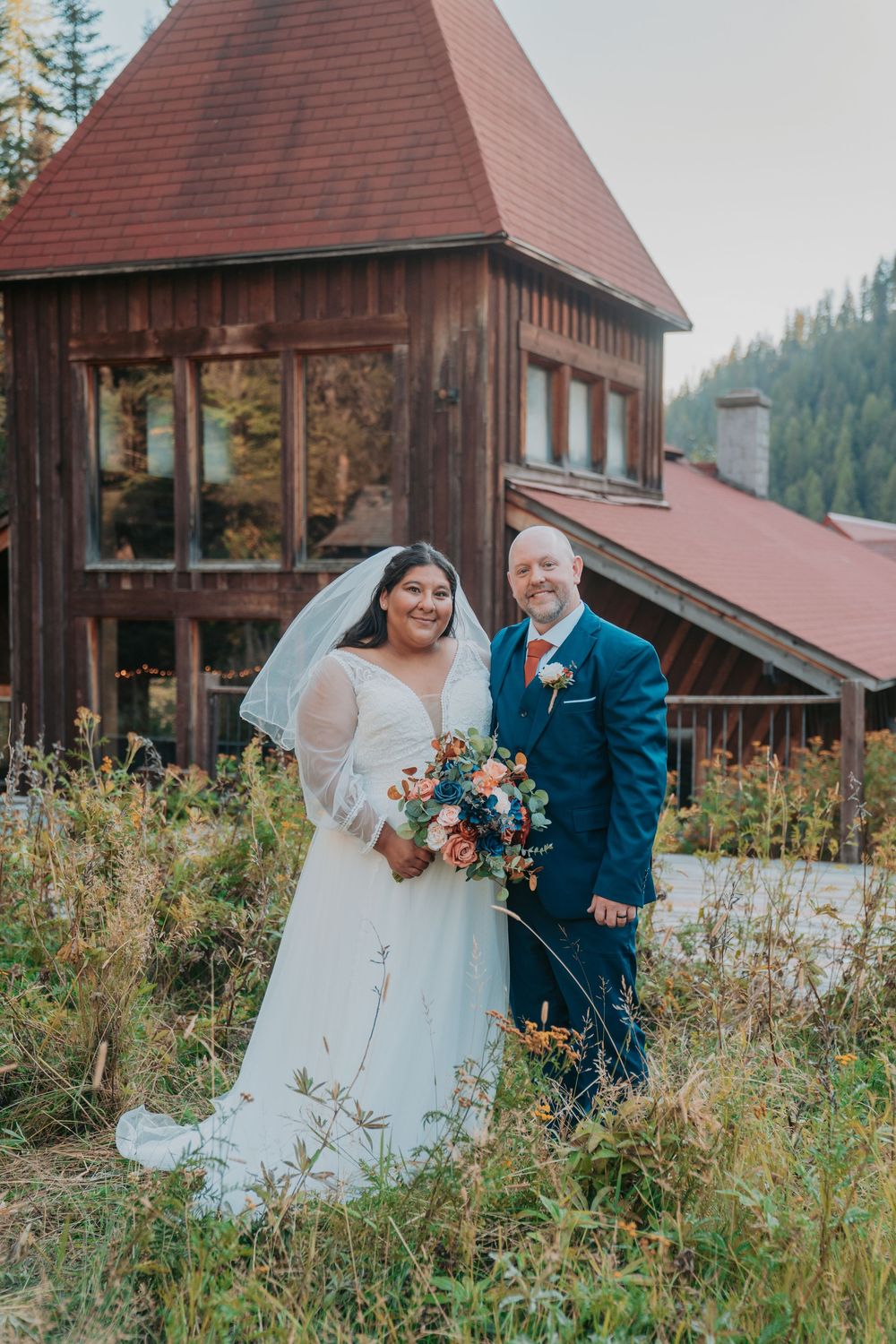 Bride and groom pose outdoors in front of a rustic wooden wedding venue surrounded by wildflowers.