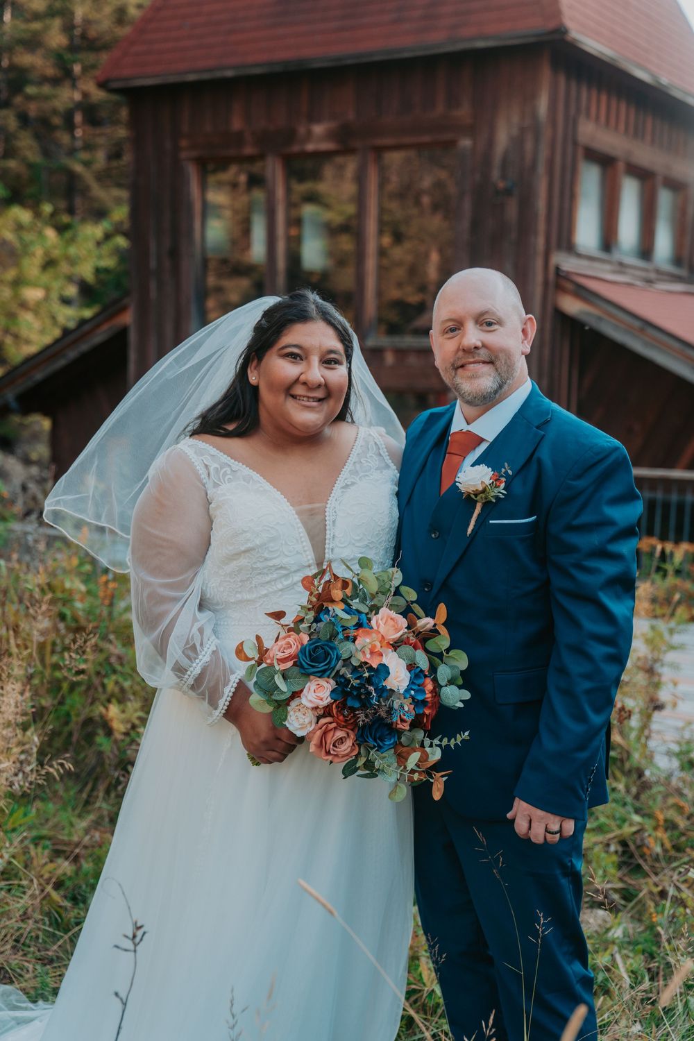 Bride in white lace gown with veil holding coral and navy bouquet stands with groom in navy suit before rustic wooden building.